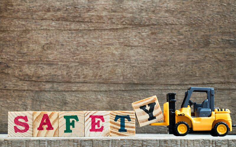 Ein Spielzeuggabelstapler hält kleine Holzwürfel, die das Wort "Safety" bilden.